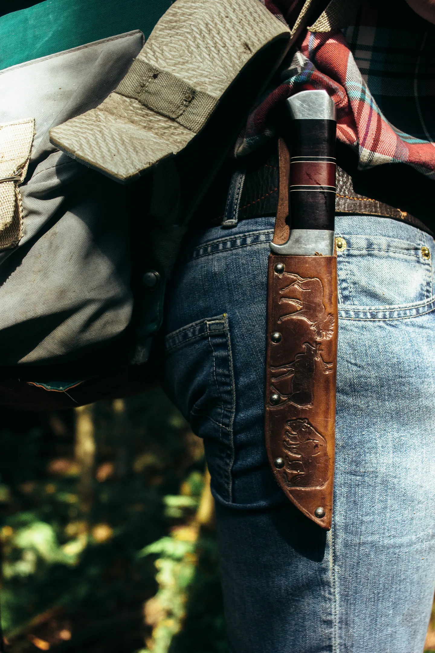 A sheathed knife worn on a belt beside a backpack in a forest.