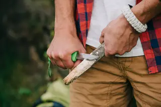 Hands carving a branch with a green-handled knife outdoors
