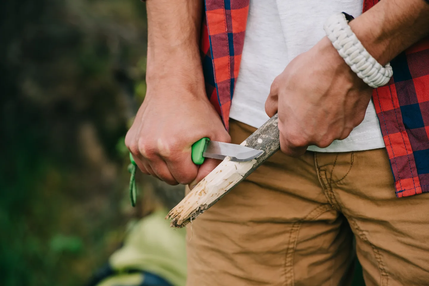 Camping knife in use: hands carving a branch with a green-handled knife outdoors
